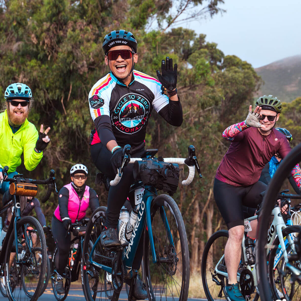 A smiling cyclist in a Cycle to Zero jersey waves to the camera while riding with a group on a tree-lined road.